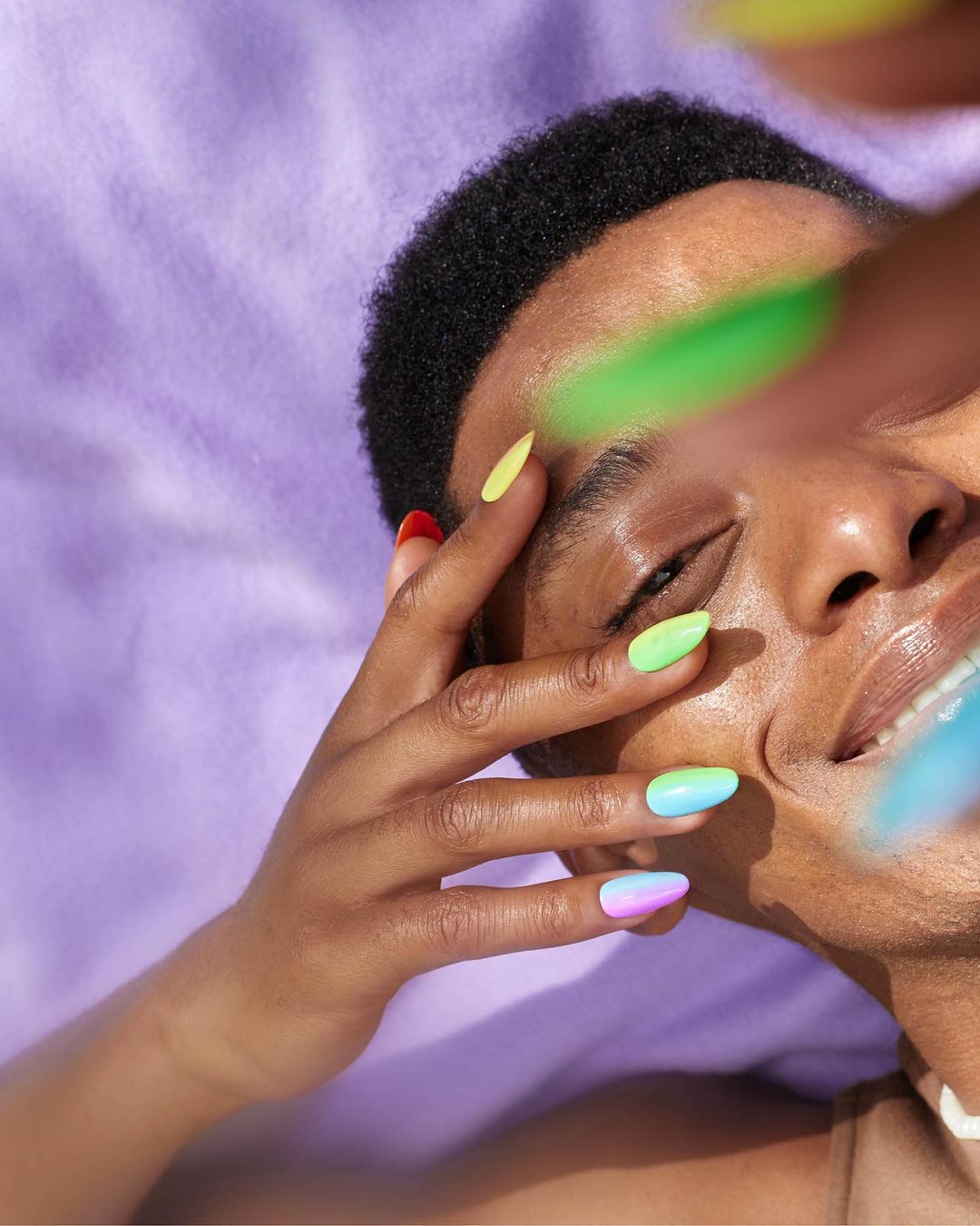 Person with multicolored nails poses against a light purple fabric backdrop, fingers near the face, smiling slightly with direct sunlight.