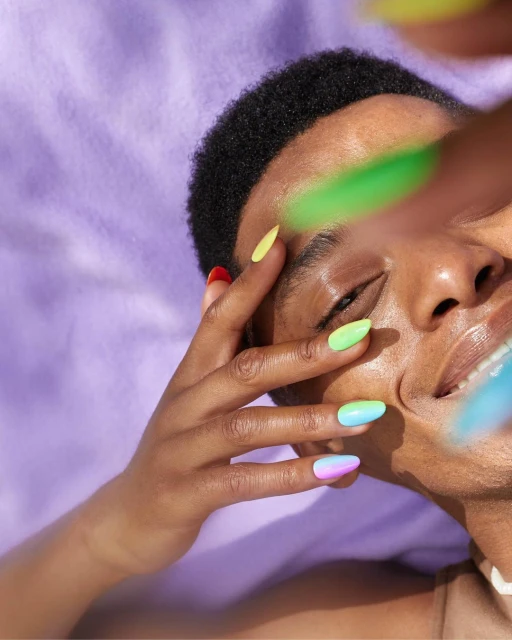Person with multicolored nails poses against a light purple fabric backdrop, fingers near the face, smiling slightly with direct sunlight.