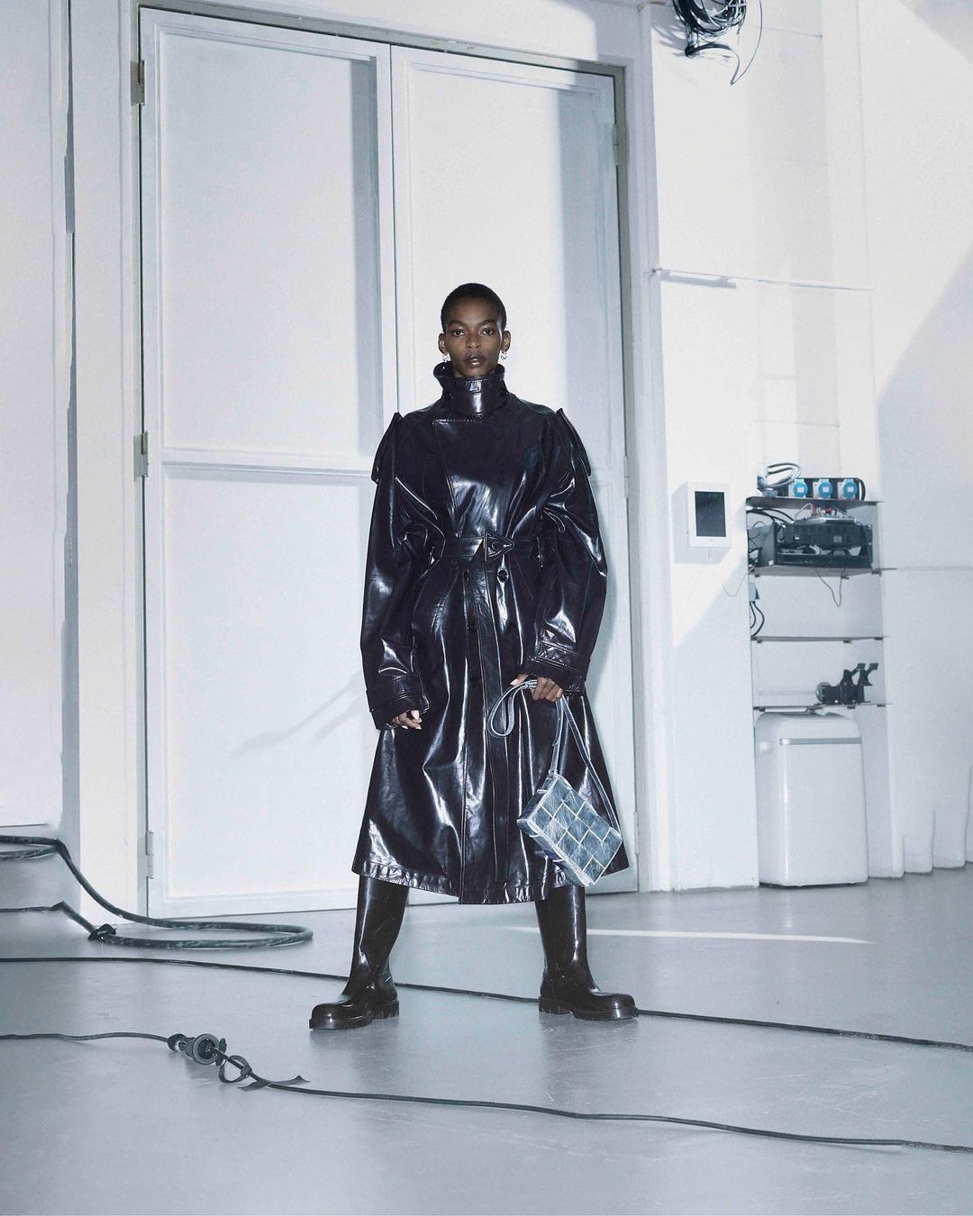 A striking fashion shot of a Black woman in a black patent-leather trench coat and boots, set against a minimalist white studio backdrop.