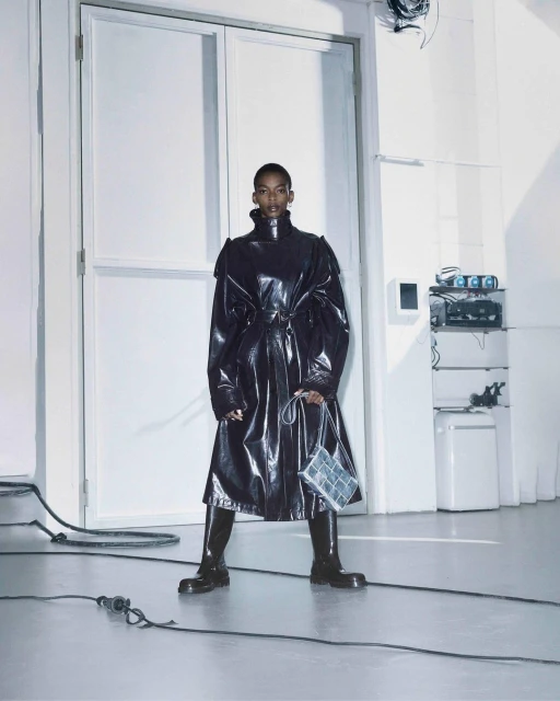 A striking fashion shot of a Black woman in a black patent-leather trench coat and boots, set against a minimalist white studio backdrop.