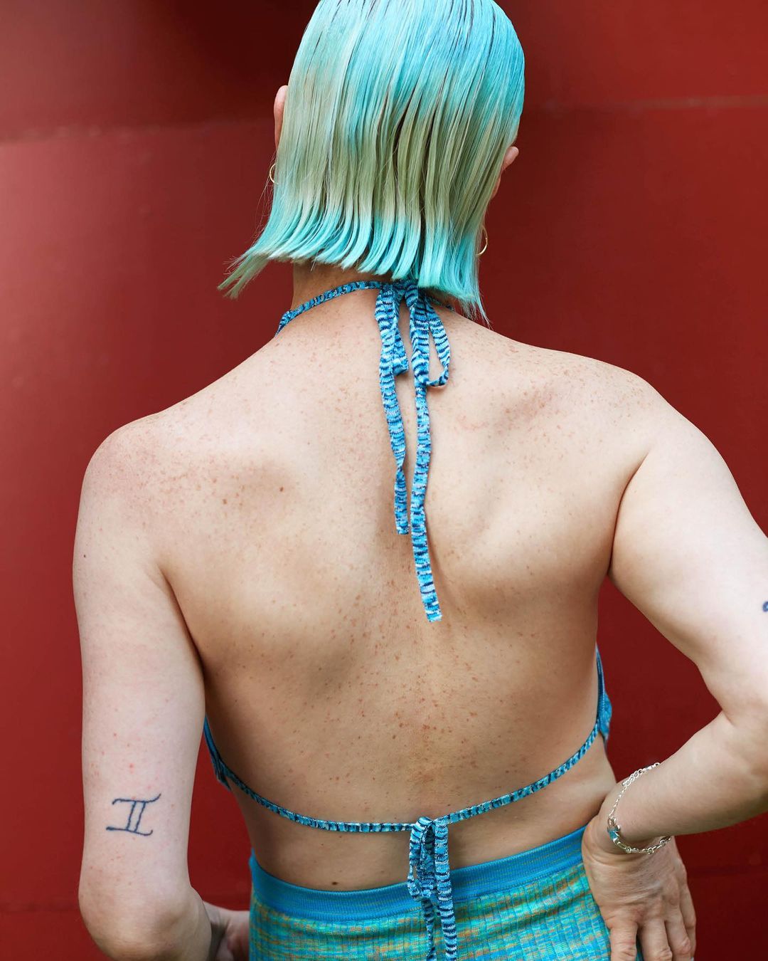 From the back, a light-skinned person with short, dyed turquoise hair wears a blue halter top and skirt against a red backdrop.