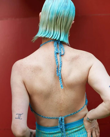 From the back, a light-skinned person with short, dyed turquoise hair wears a blue halter top and skirt against a red backdrop.