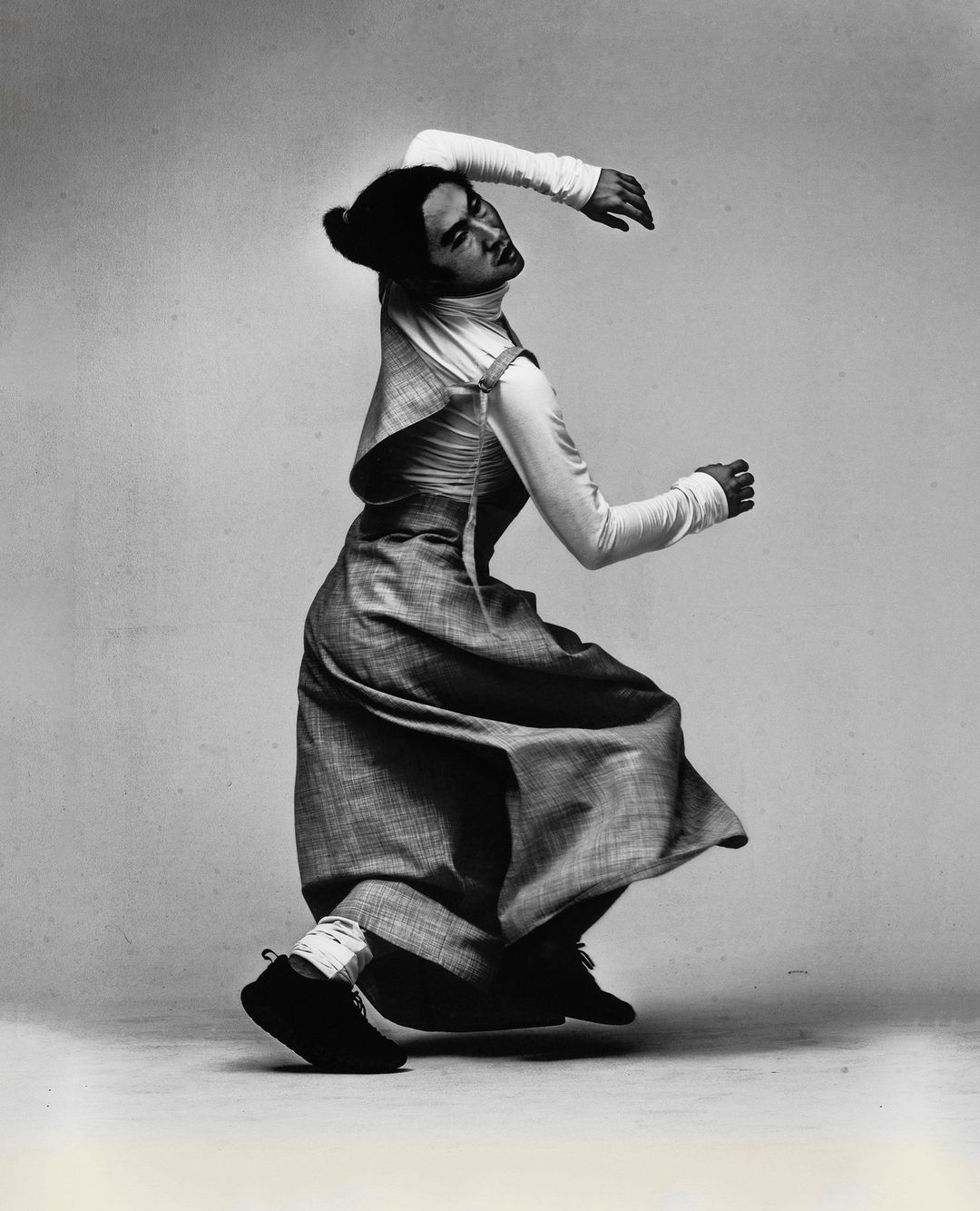 Monochrome studio shot of an adult female in mid-motion, wearing a dress over a long-sleeve shirt and sneakers, against a plain backdrop.