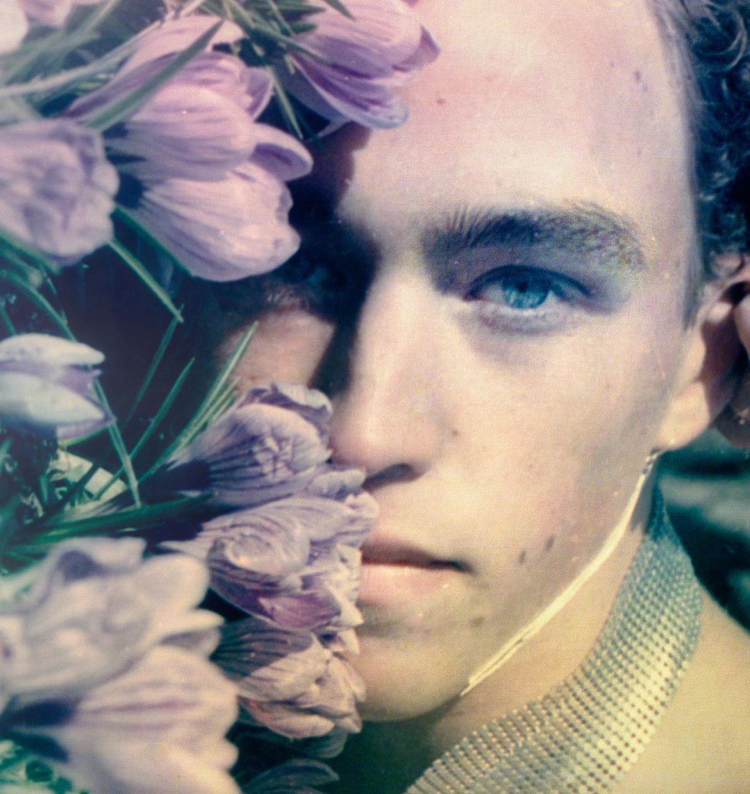 Close-up portrait of a young adult with fair skin, blue eyes, and curly brown hair, adorned with purple flowers and a metallic garment.