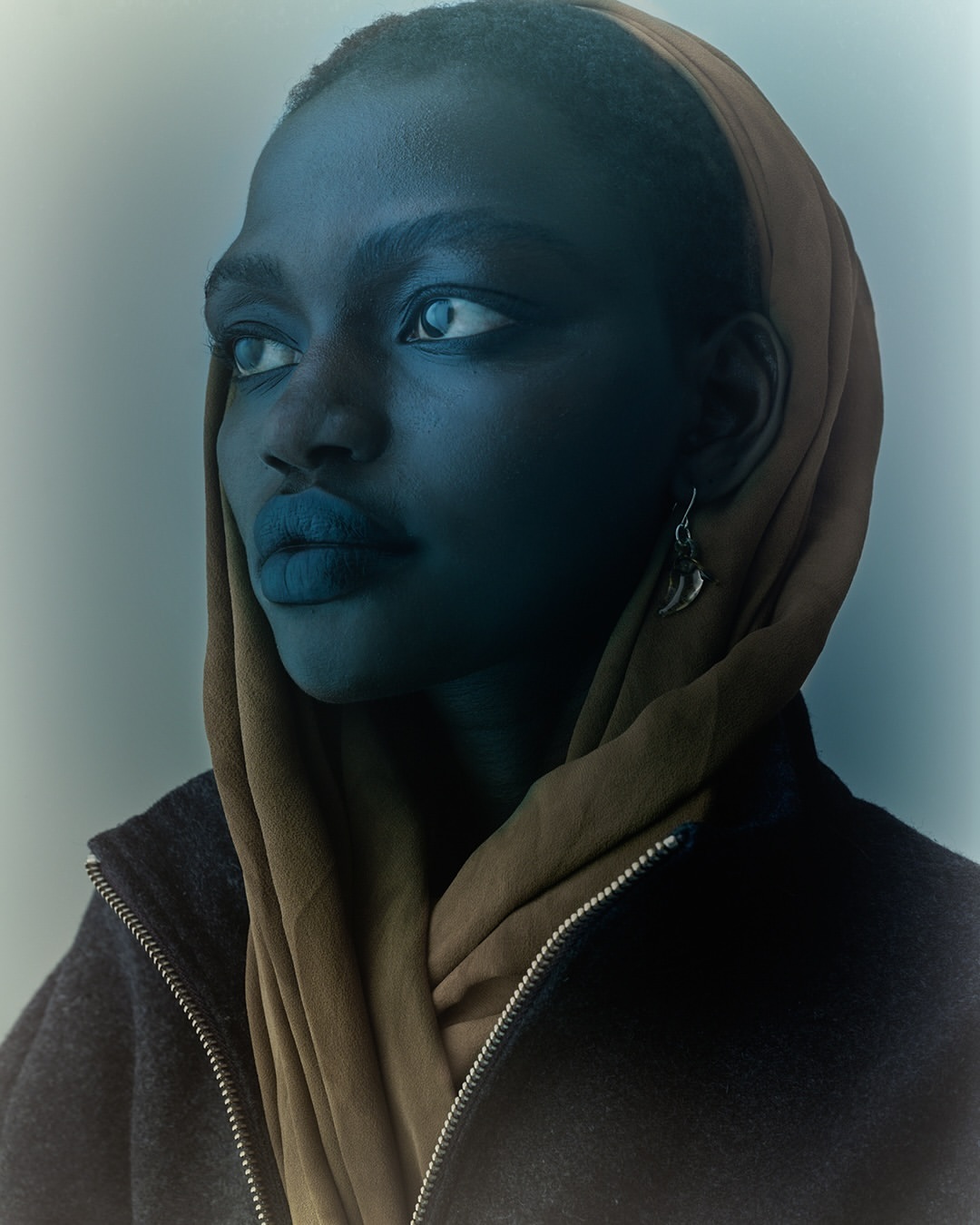 Portrait of a Black female model with a brown headscarf and dark jacket, looking away from the camera in a soft, diffused light.