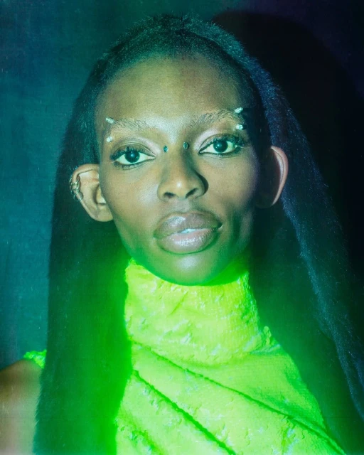 Close-up portrait of a woman with dark skin, light-colored eyebrows and decorations, wearing a neon-green dress against a dark background.
