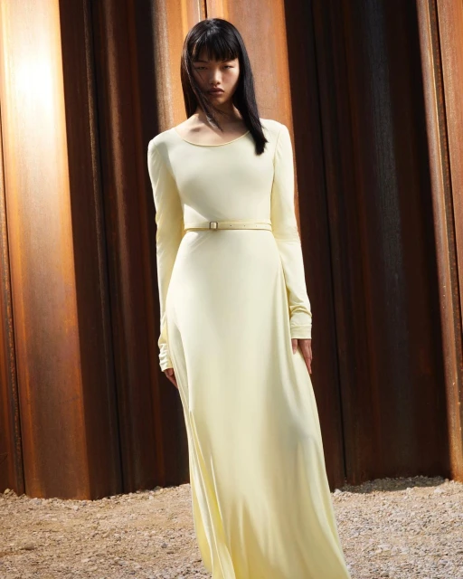 Asian woman in long-sleeved yellow maxi dress with belt, standing against a rust-colored textured metal backdrop, fashion editorial.