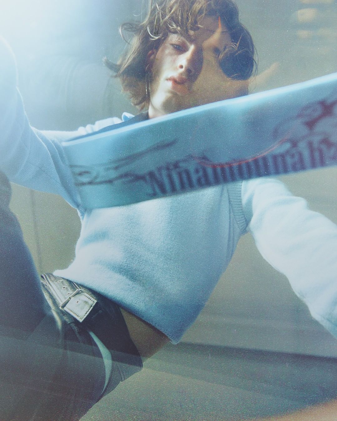 Close-up shot of a young adult male model with long brown hair, wearing a light blue sweater vest and jeans, posing indoors with bright lighting.