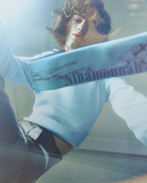 Close-up shot of a young adult male model with long brown hair, wearing a light blue sweater vest and jeans, posing indoors with bright lighting.