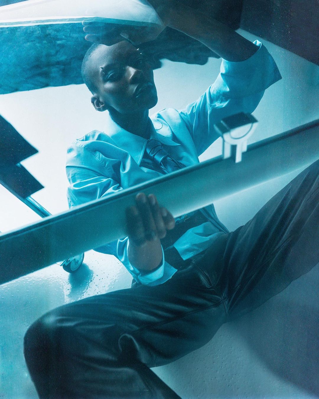 An adult with short hair wearing a blue shirt, tie, and dark pants poses in a studio with an abstract, geometric backdrop.