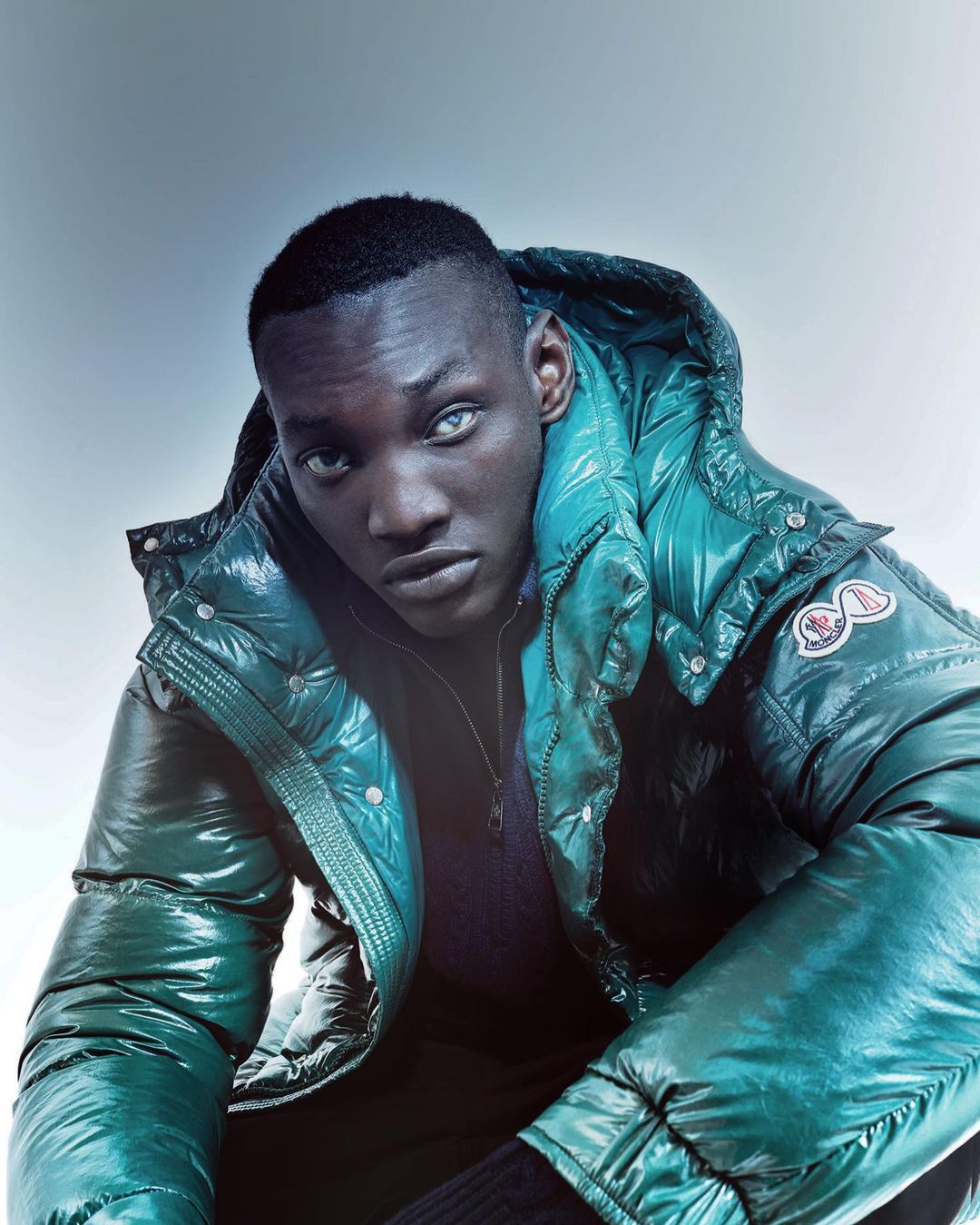 Fashion portrait of an adult Black male model wearing a shiny green puffer jacket, medium studio shot, pale background, neutral expression.