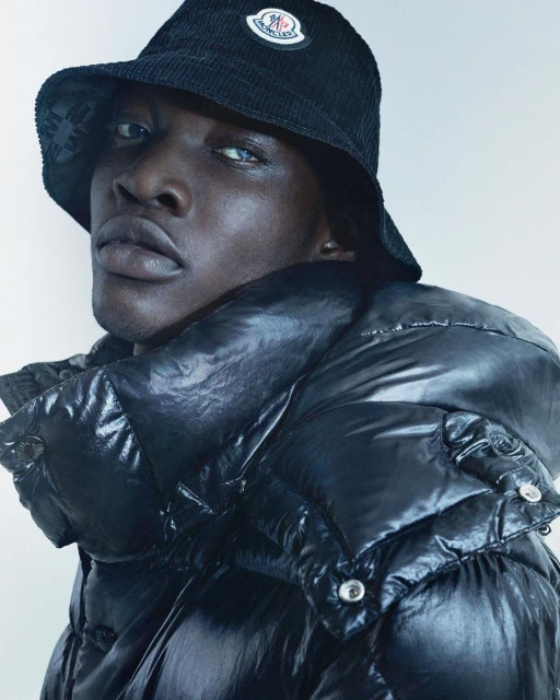 Portrait of an adult male wearing a black corduroy bucket hat with a Moncler logo and a black shiny puffer jacket indoors.