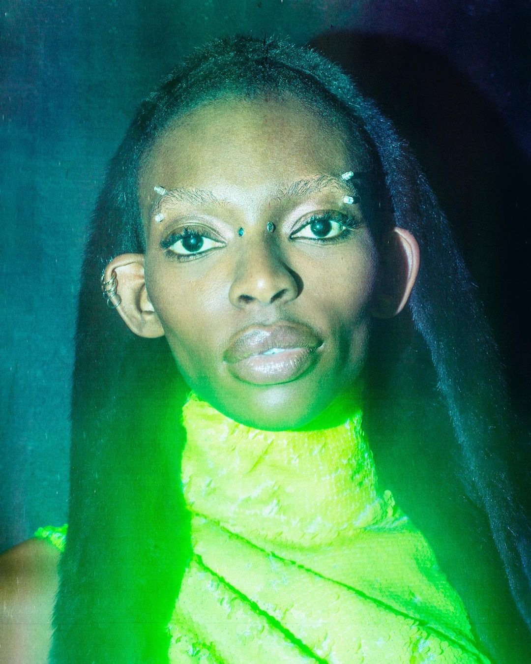 Portrait of a Black woman with dark hair and beaded eyebrows wearing a bright yellow top against a dark, moody background.
