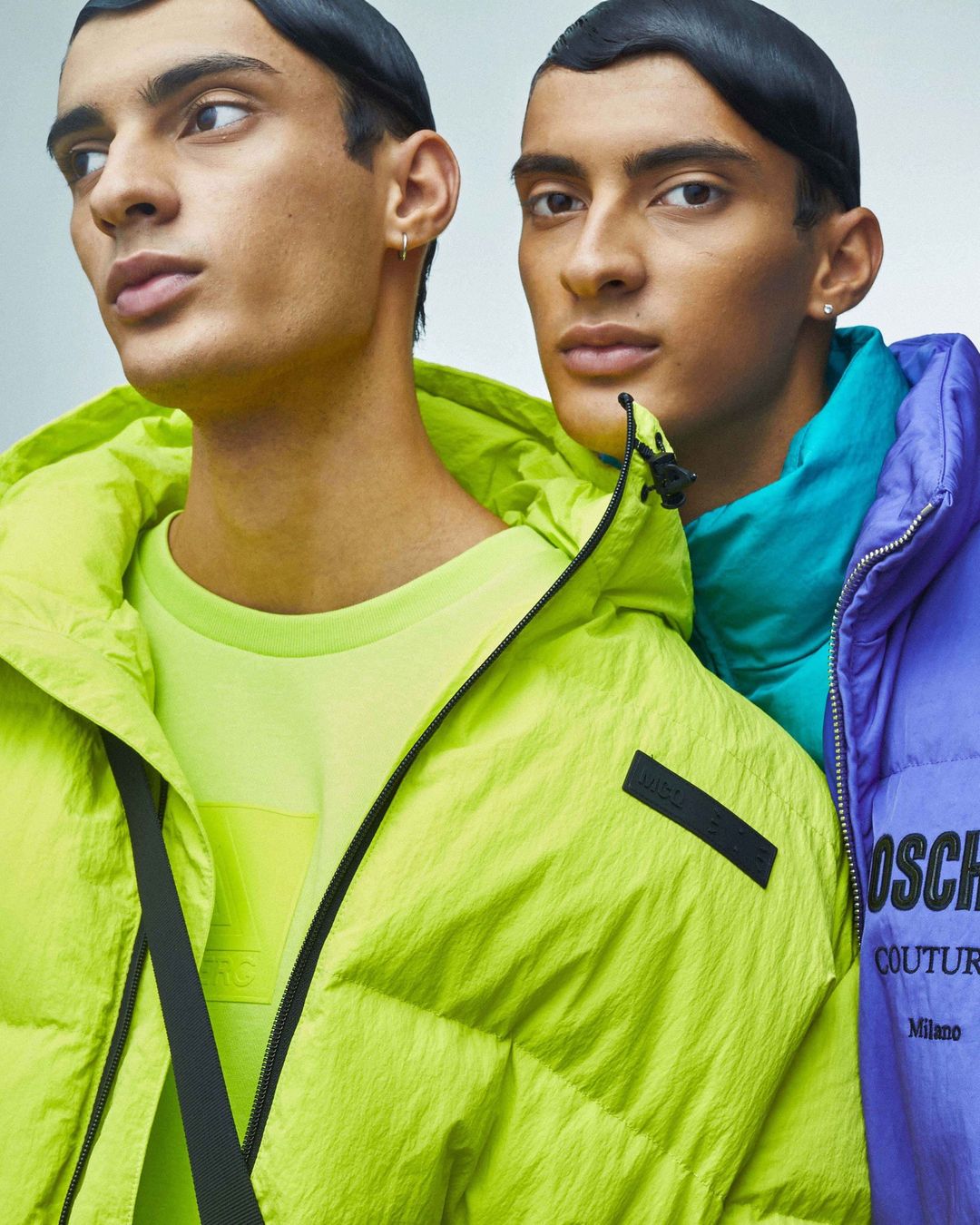 Two adult male fashion models pose indoors in neon jackets; one in yellow, the other in purple and teal, against a gray background.