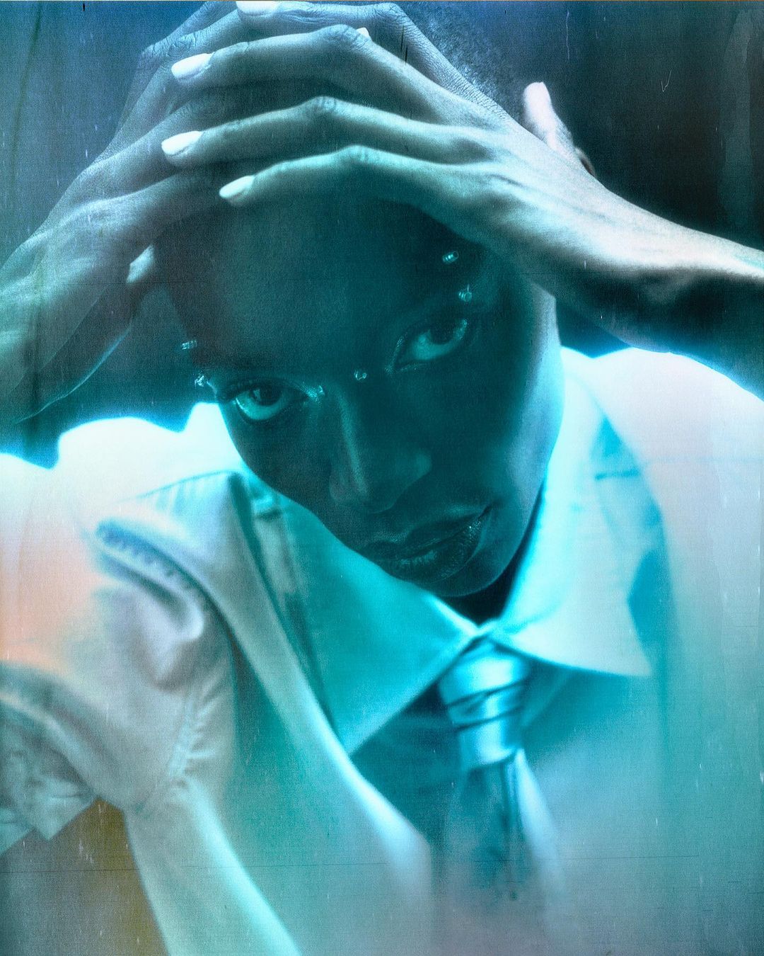 Close-up portrait of an adult woman with her hands on her head, wearing a white collared shirt and blue tie, under blue lighting.