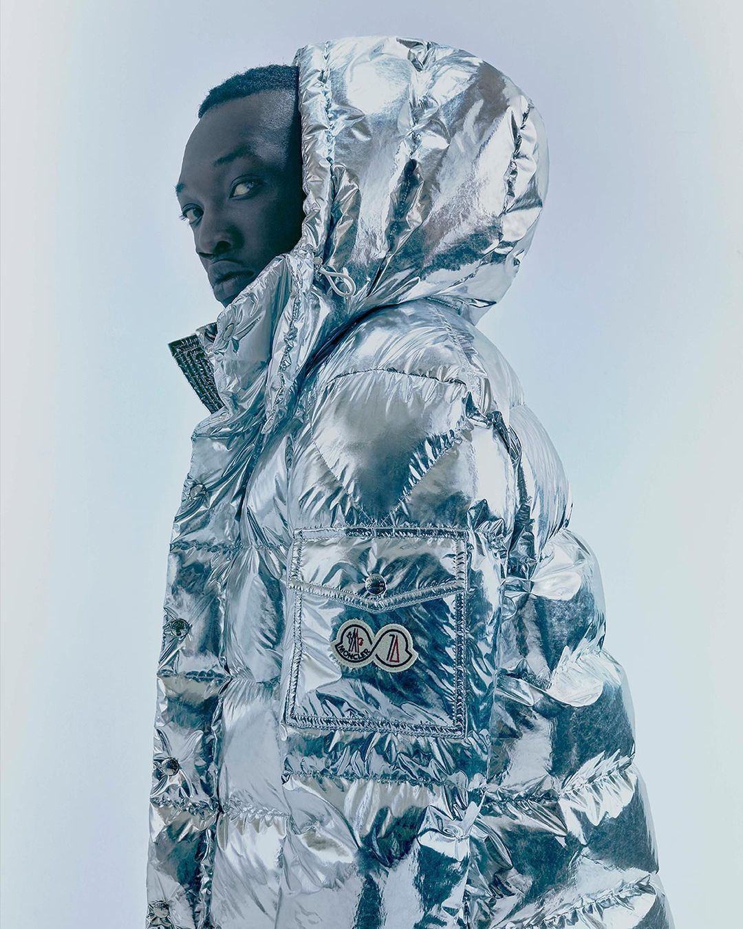Stylish portrait of a Black man in a shiny, silver hooded puffer jacket against a bright backdrop, showcasing modern fashion.