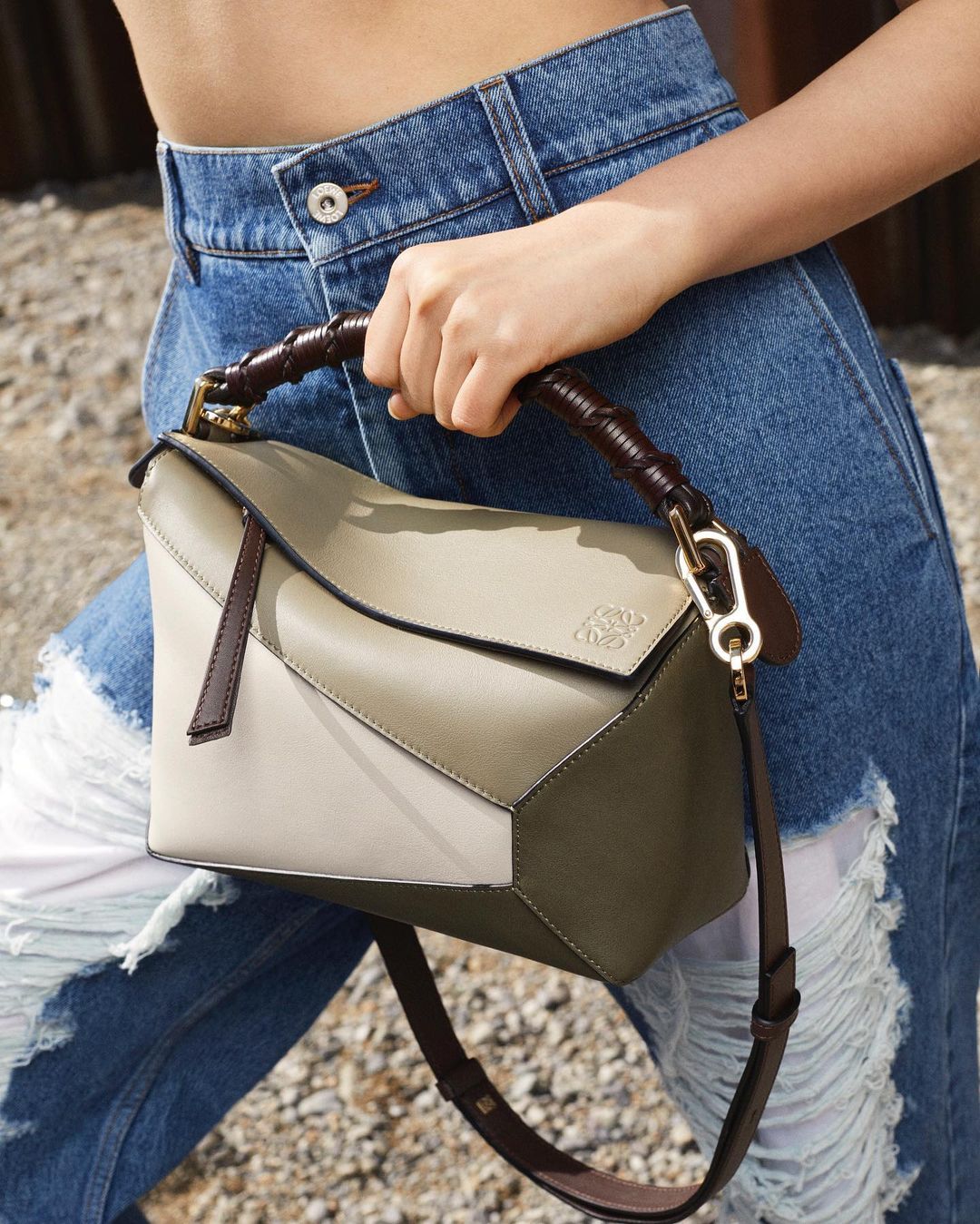 Tan and green Loewe Puzzle bag with leather strap and bamboo handle, worn with light blue ripped jeans, against a neutral background.