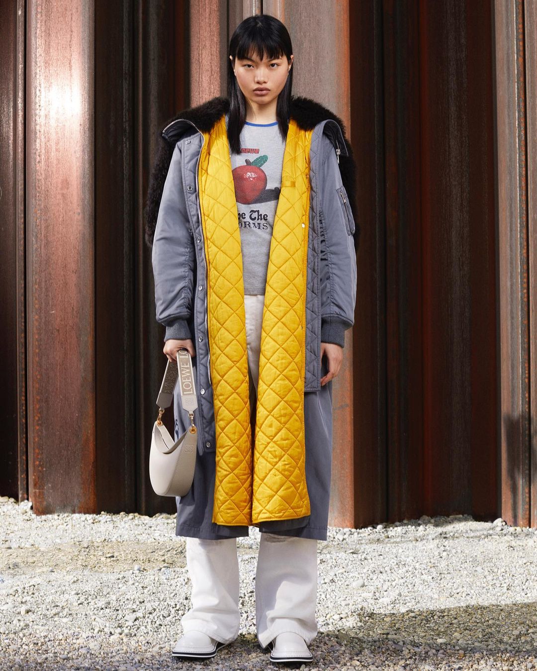 A woman stands in an outdoor setting, wearing a long coat with a yellow scarf, cream pants and holding a cream colored purse.
