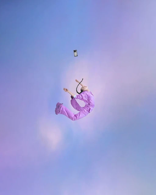 Person wearing lilac jumpsuit jumps as a smartphone floats overhead against a light blue and violet cloudy backdrop in studio setting.
