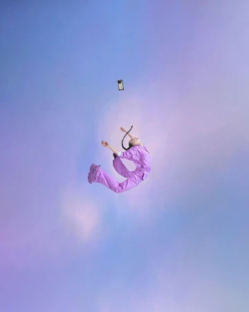 Person wearing lilac jumpsuit jumps as a smartphone floats overhead against a light blue and violet cloudy backdrop in studio setting.