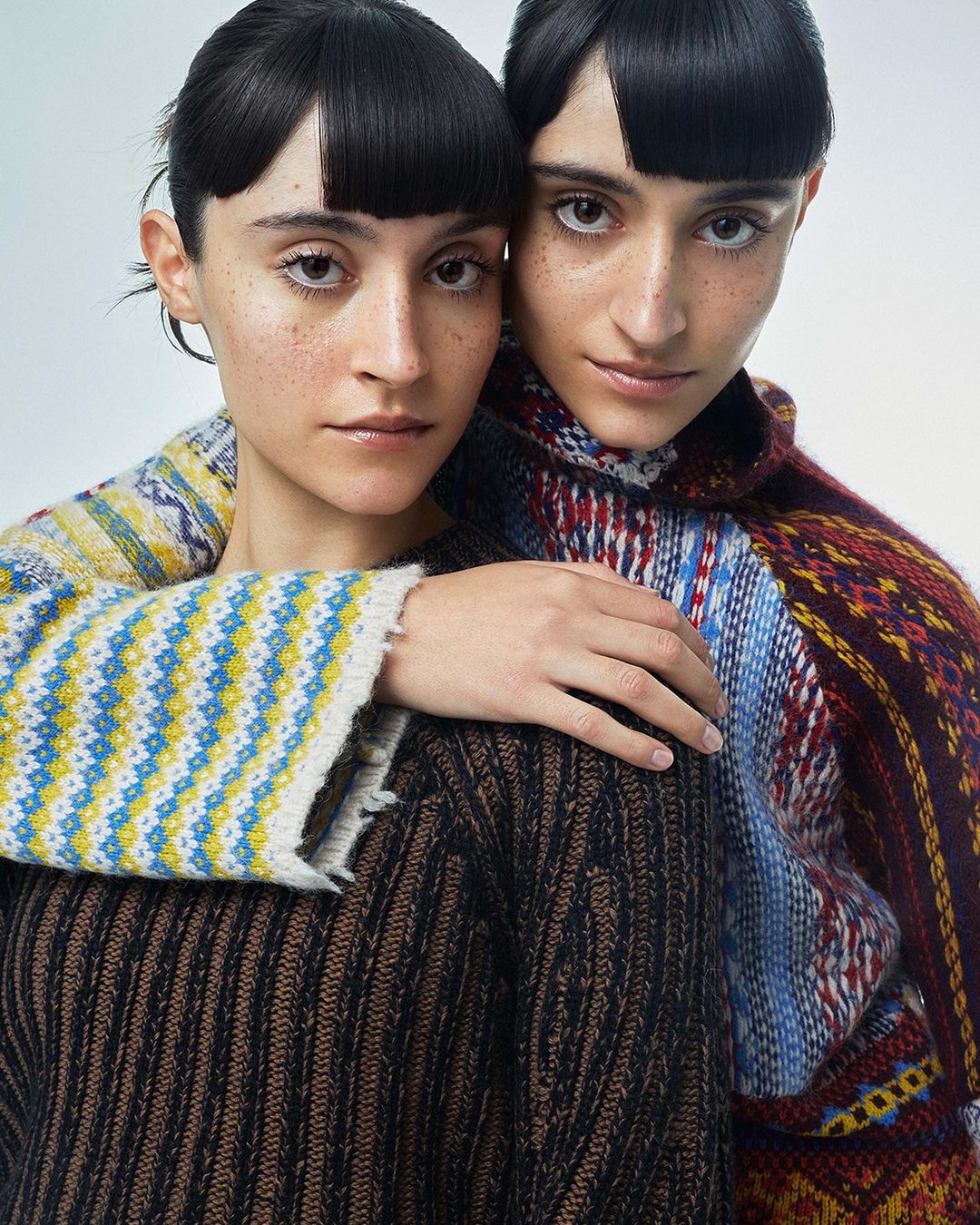 Two women with matching haircuts and knitted sweaters pose closely together against a neutral backdrop in an intimate portrait shot.