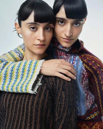Two women with matching haircuts and knitted sweaters pose closely together against a neutral backdrop in an intimate portrait shot.