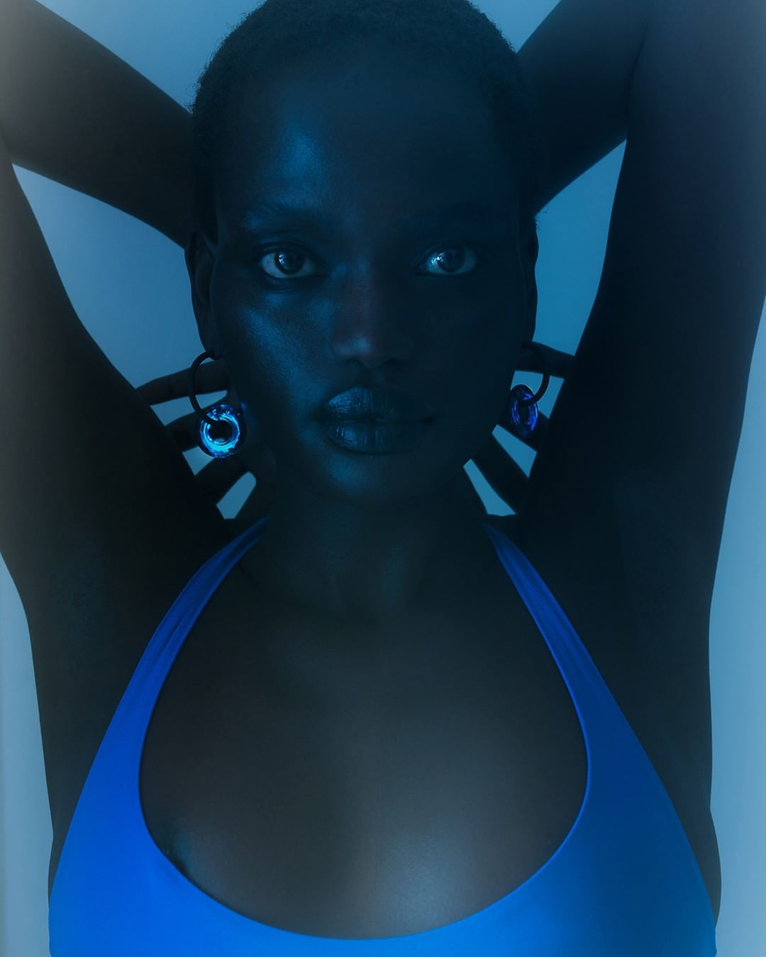Portrait of a woman with dark skin and blue top, looking at camera with hands behind her head, set against a blue backdrop.