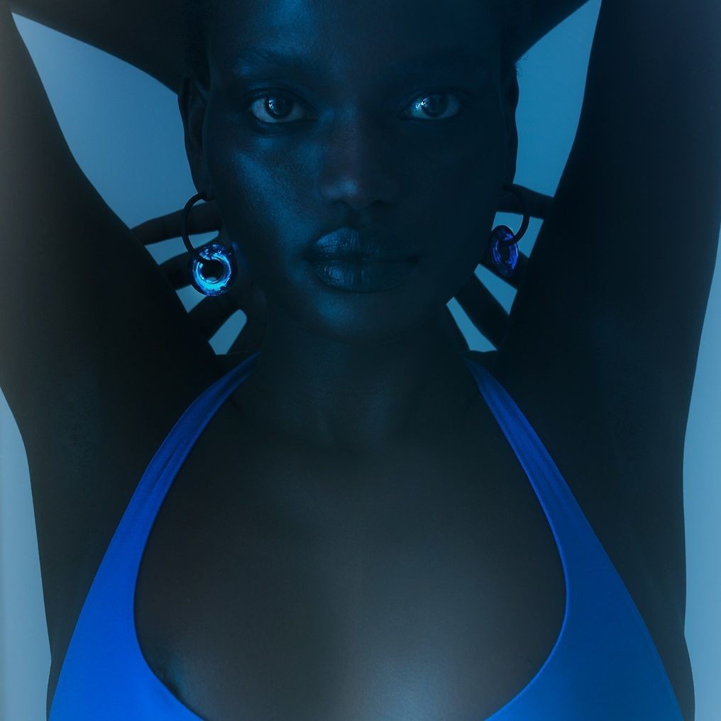 Close-up of a dark-skinned woman in blue swimsuit, wearing circular earrings and posing with hands behind her head against a blue backdrop.