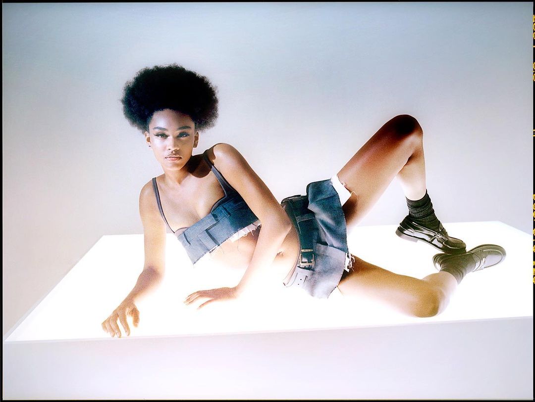 Young woman with Afro poses on a bright white surface wearing a denim top and skirt with black shoes and socks in a studio setting.