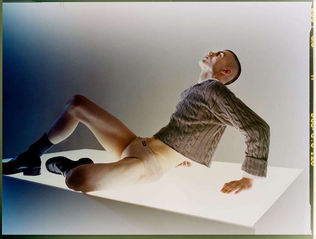 An adult man poses in a tan sweater, underwear, and black shoes, reclining on a white surface against a gradient background.