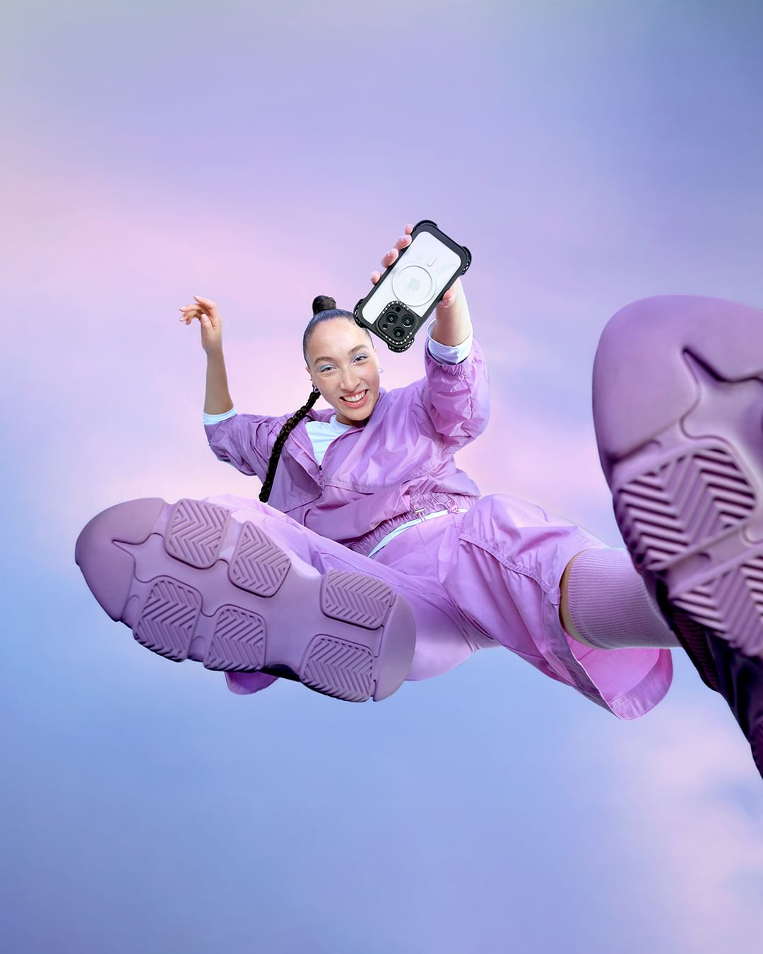 A woman in a lavender tracksuit leaps toward the camera holding her phone with a protective case with pastel purple sky background.