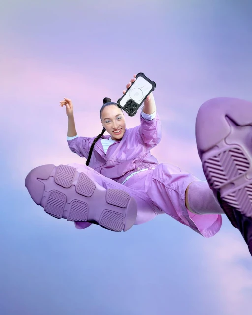 A woman in a lavender tracksuit leaps toward the camera holding her phone with a protective case with pastel purple sky background.