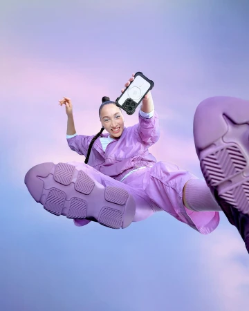 A woman in a lavender tracksuit leaps toward the camera holding her phone with a protective case with pastel purple sky background.