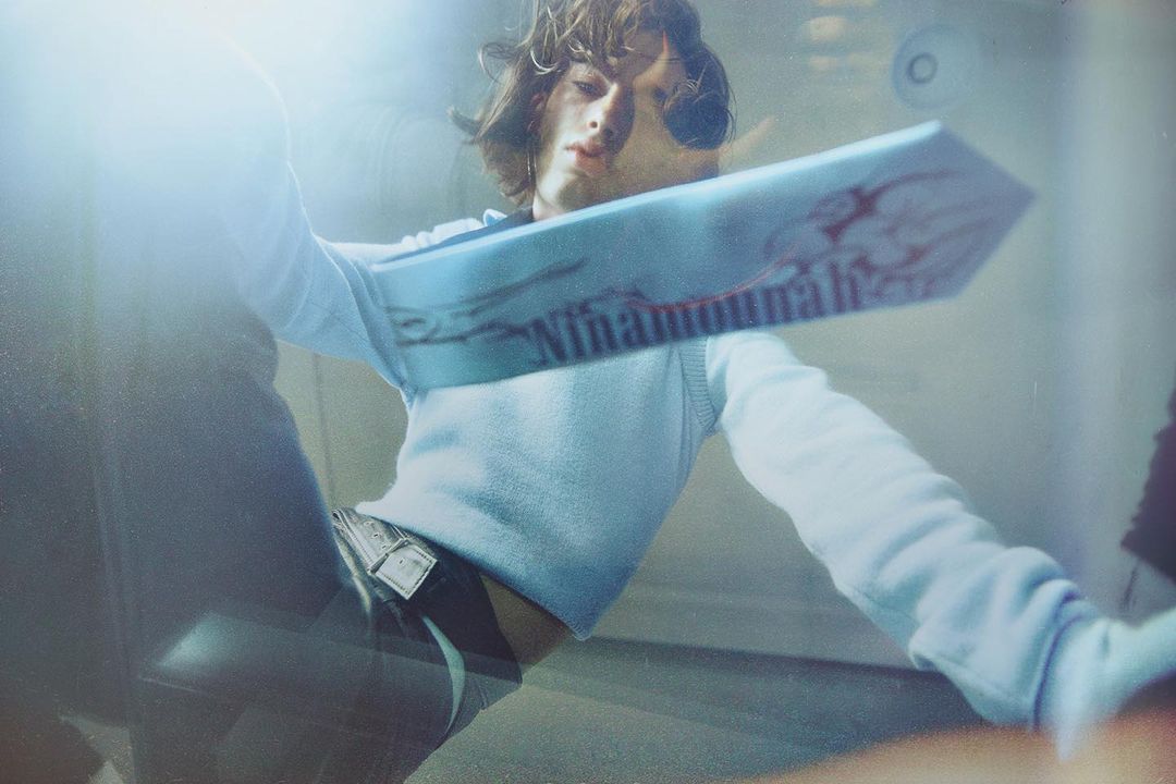 Indoor shot of a person with long brown hair wearing a blue sweater and jeans, holding a blue object with text; bright lighting.