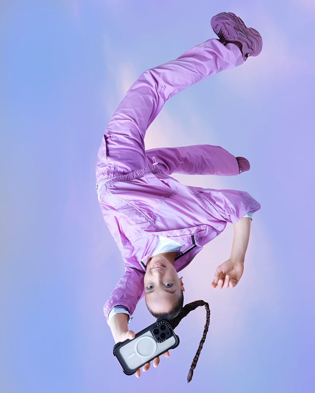 Young woman in athletic gear, posing upside down, showcases phone. Fashion-forward athletic wear, ponytail, against soft pastel backdrop.