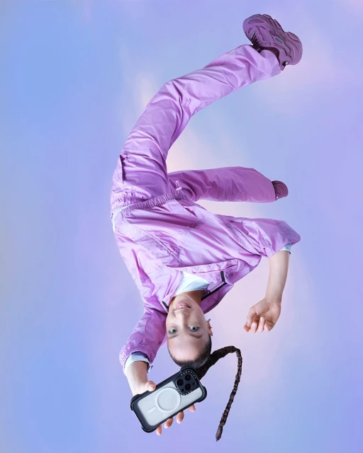Young woman in athletic gear, posing upside down, showcases phone. Fashion-forward athletic wear, ponytail, against soft pastel backdrop.