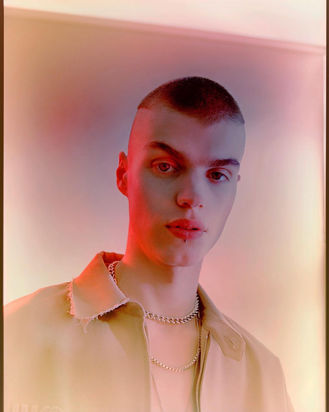 Portrait of a young adult male with a shaved head wearing a tan collared shirt and silver chain necklaces, with pink and yellow lighting.