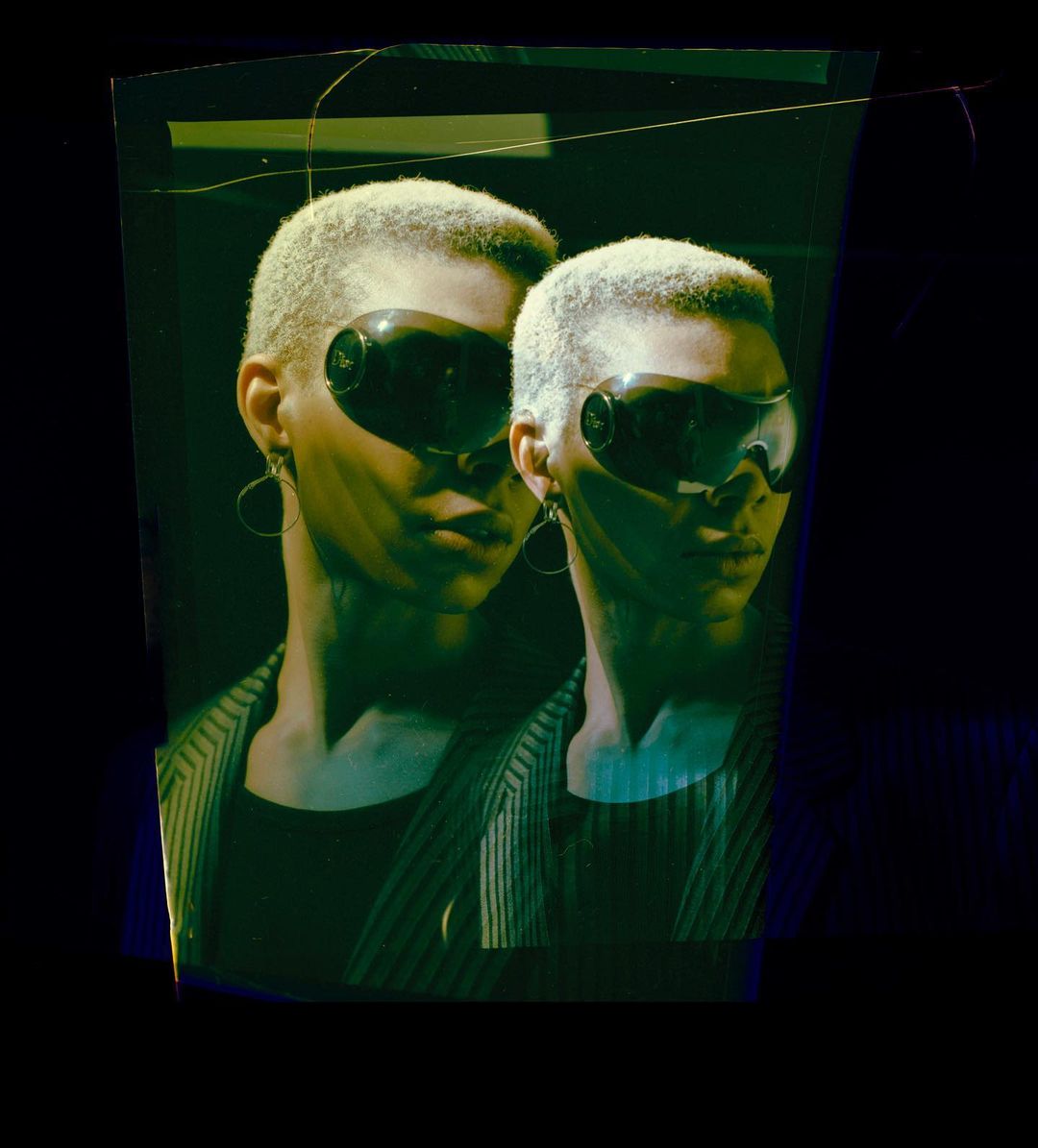 Two adult females pose indoors with sunglasses, wearing striped blazers with short hair, in a close-up, high-contrast studio fashion shot.