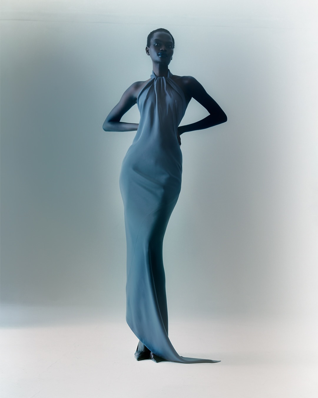 Fashion shot of a Black woman posing in a light blue halter gown, hands on hips, against a pale blue studio backdrop with soft lighting.