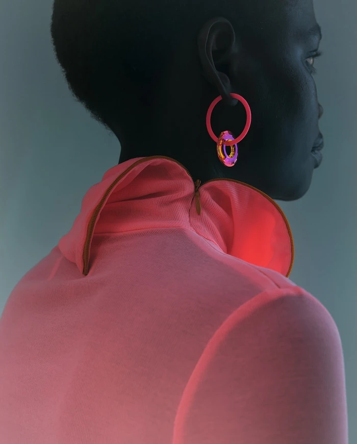 Close-up profile of a black woman wearing a pink turtleneck and bright pink hoop earrings against a grey background.
