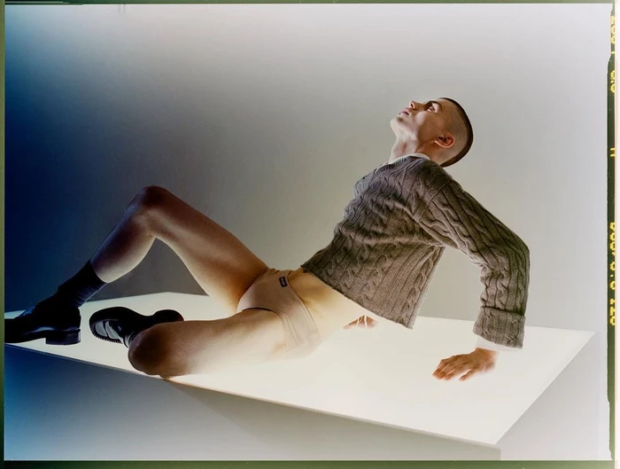 Man with a shaved head wearing a cable knit sweater and underwear leaning back on a glowing white platform