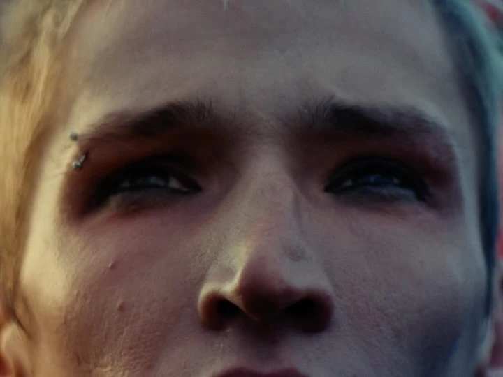 Close-up on face of young adult with blond hair, a piercing on their left eyebrow, and an upward gaze; natural lighting, muted color palette.