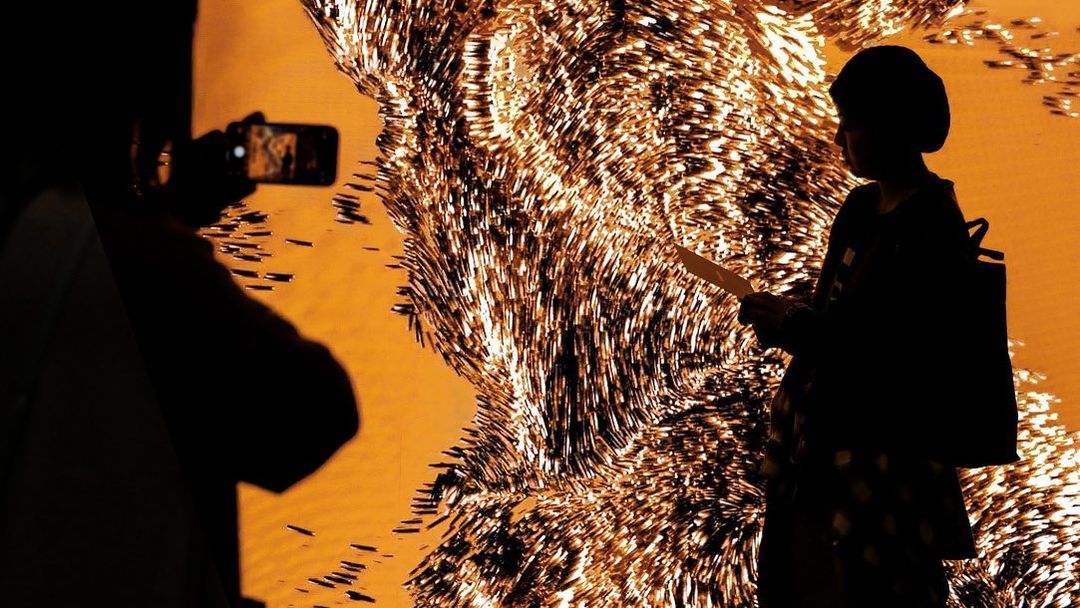 Silhouetted figures interact with a shimmering art installation in shades of gold and black, capturing a moment of artistic appreciation.