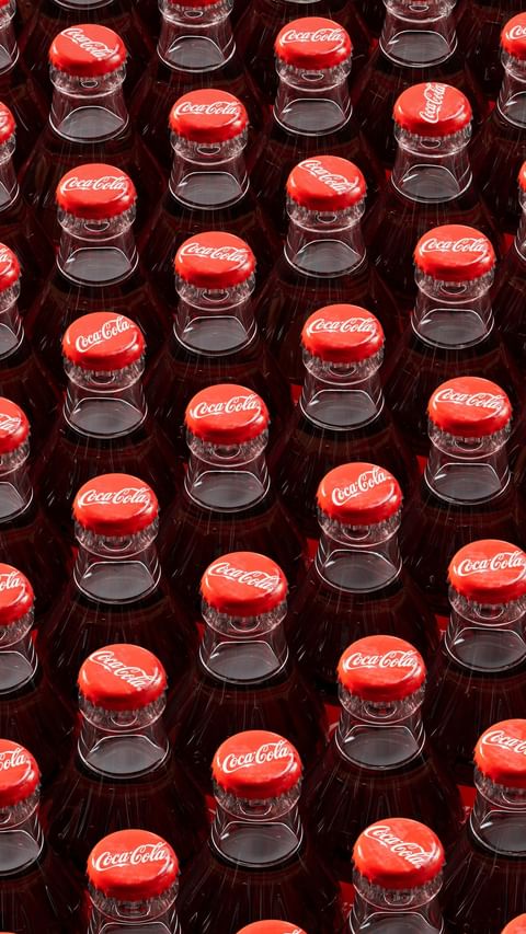 High-angle, full-frame shot of multiple Coca-Cola bottles with red caps and logo. The bottles are densely packed, creating a repeating pattern.