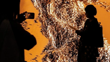 Silhouettes of museum visitors viewing a large glowing textured art installation with warm orange lighting