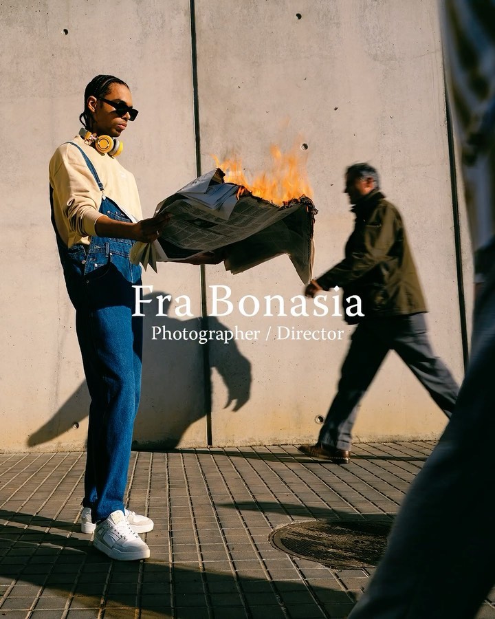 Young man in overalls holds burning newspaper on city street. Another man walks in the background, captured at medium shot. Concrete wall backdrop.