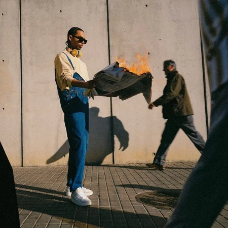 Young adult holds burning newspaper, backlight, street scene. Pedestrian in background, urban environment, city sidewalk, daytime. 