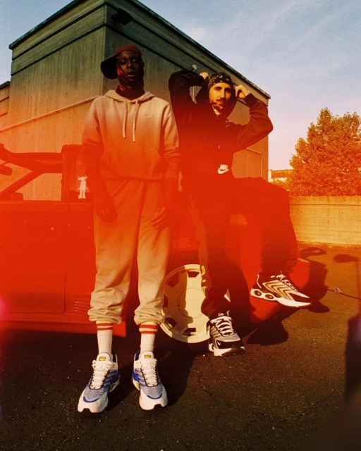 Two men pose in front of a vintage red vehicle on a sunny day, wearing casual athletic wear and colorful sneakers. The backdrop features an urban setting.