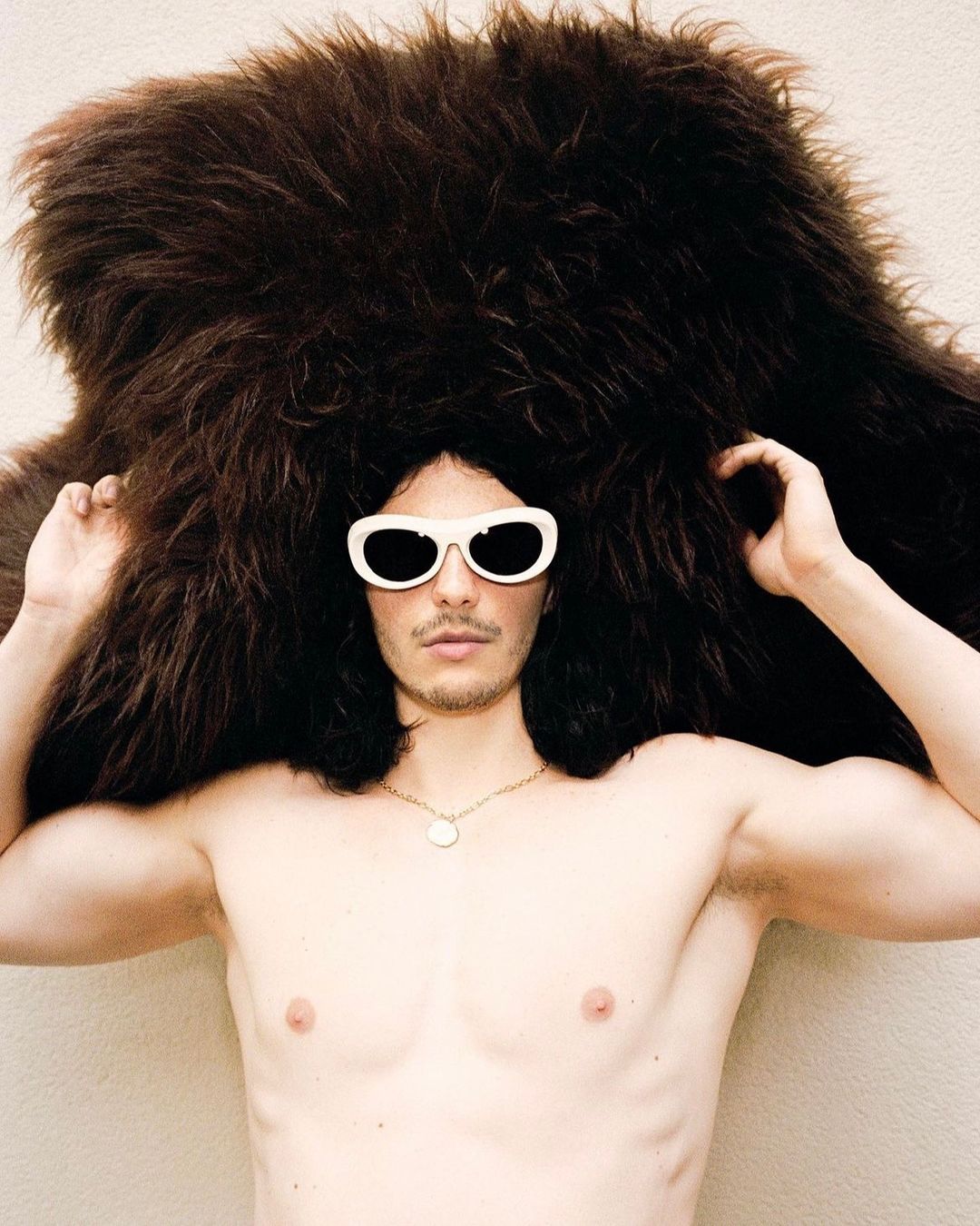 Shirtless man wearing sunglasses and a furry hat posing indoors. The man has dark hair and is wearing a gold chain necklace.