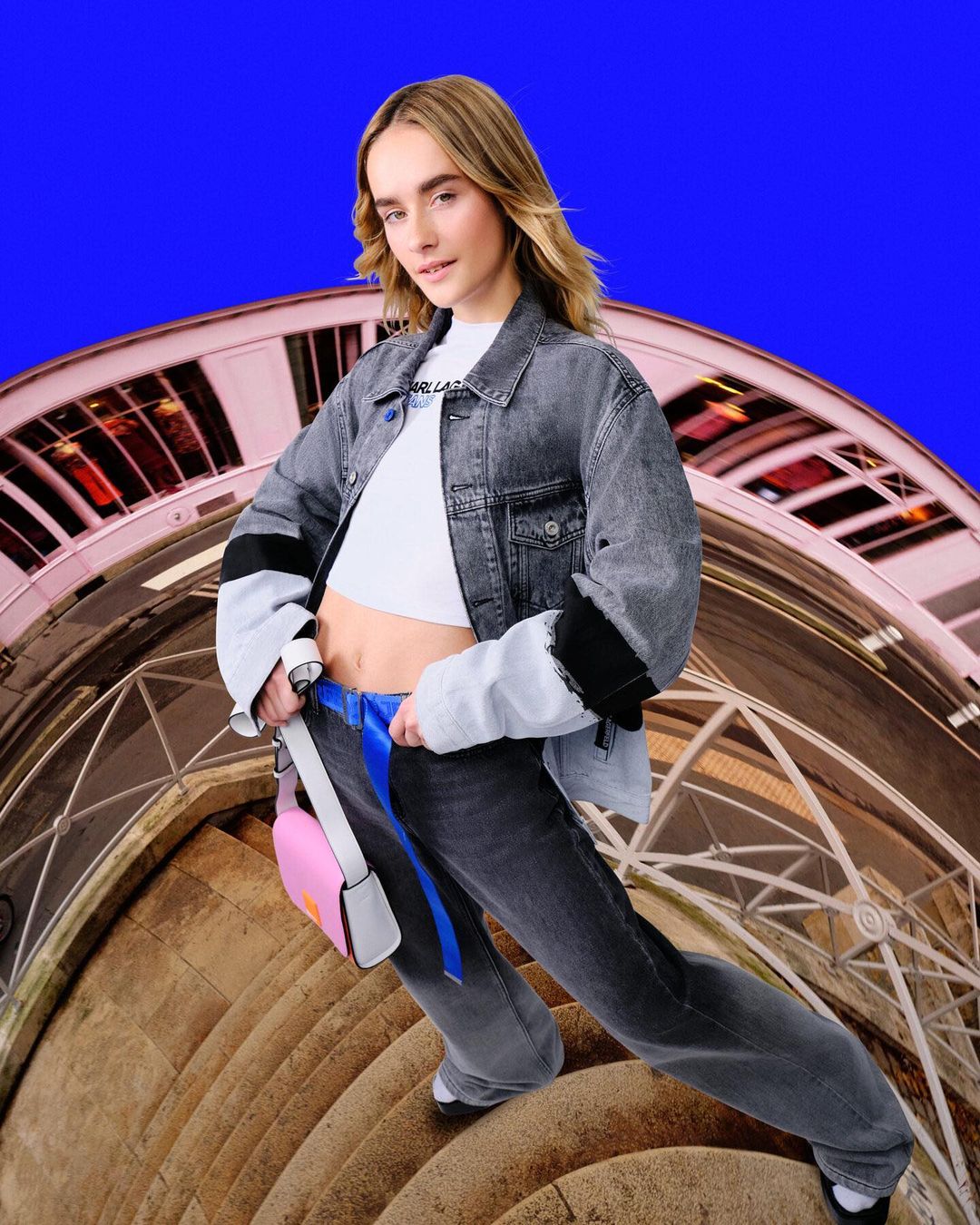 A young adult woman poses in a gray denim jacket and matching jeans with a pink purse on a stone staircase against a vibrant blue sky backdrop.